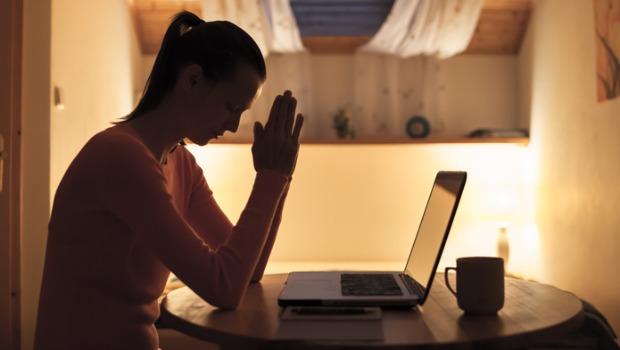 A woman sits and prays in front of her laptop.